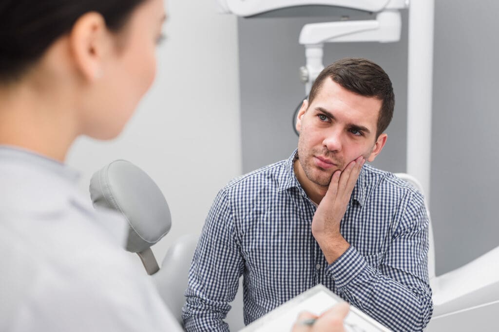 Male patient in checkered shirt displays facial pain in dental chair; dentist in East Lansing assesses and records symptoms.