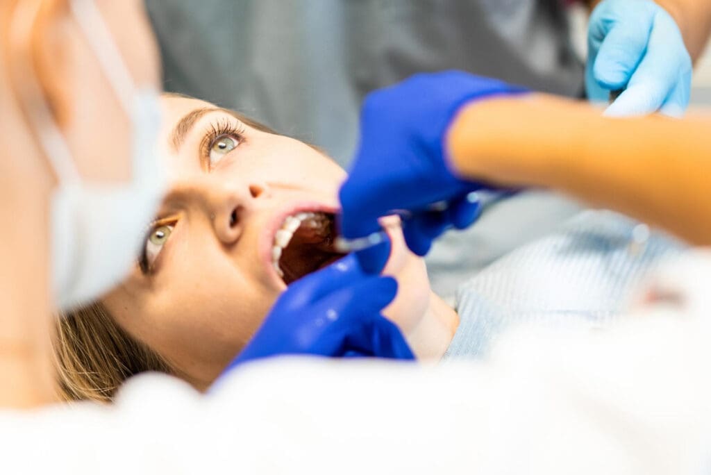 Dentist in East Lansing, MI, with blue gloves examines patient's teeth using standard tools; partially visible face in bright operatory.