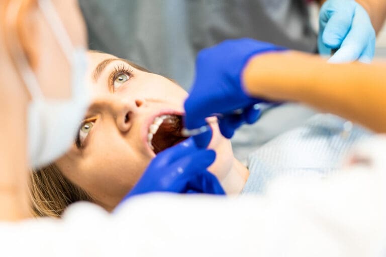 Dentist in East Lansing, MI, with blue gloves examines patient's teeth using standard tools; partially visible face in bright operatory.