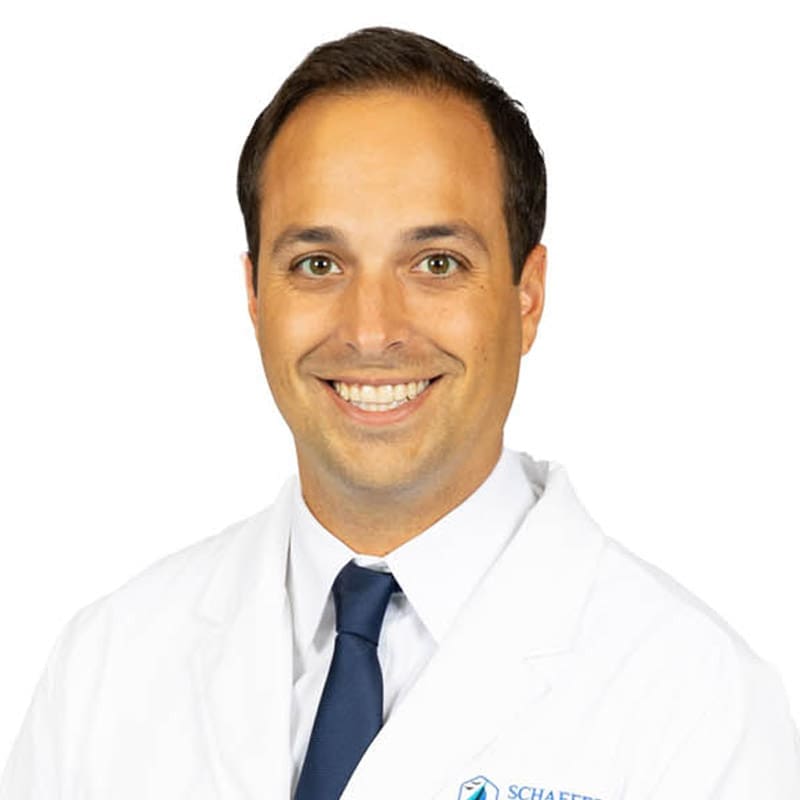 Smiling male dentist in East Lansing, MI, with short brown hair, lab coat, shirt, and blue tie stands against white background.